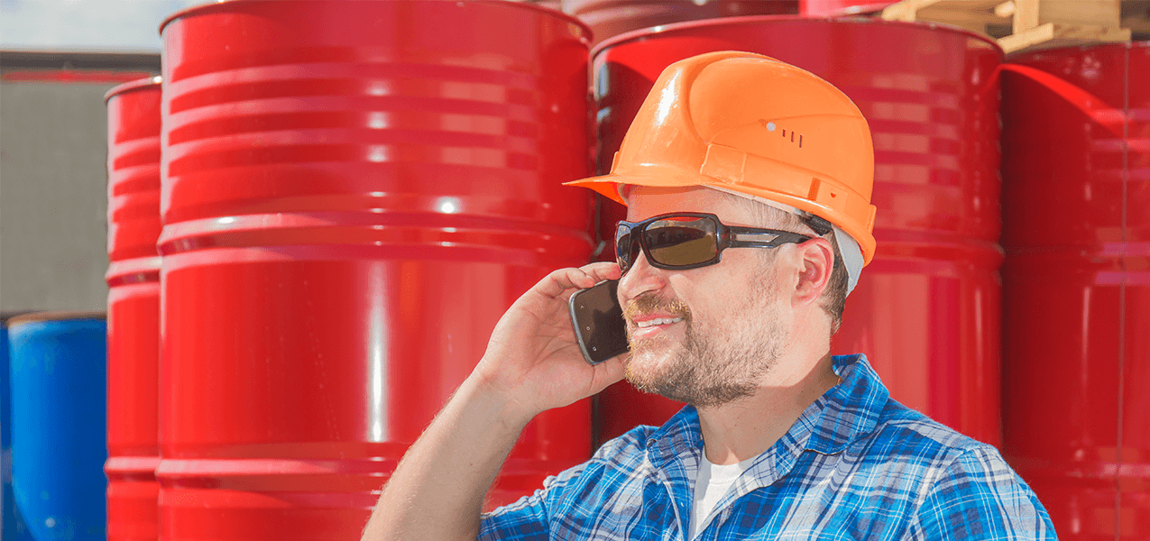 Mann mit Sonnenbrille und orangefarbenem Schutzhelm telefoniert vor mehreren roten Fässern.